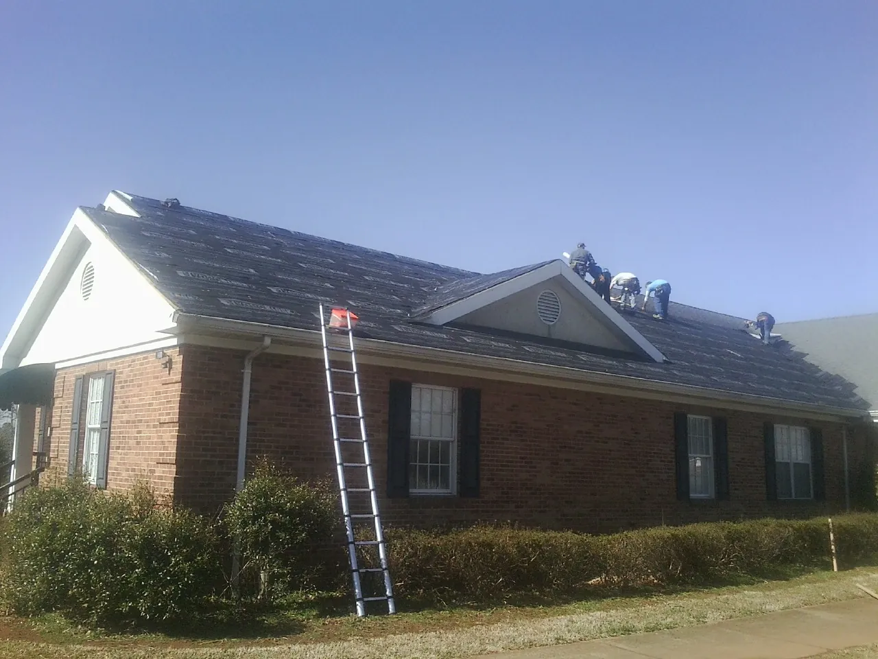Crew tearing shingles off a brick ranch roof with a ladder set — Metal Roof Repair work by our crew in Pease